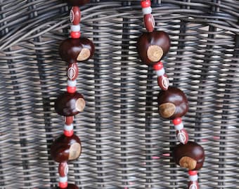 May include: A handmade necklace featuring large, dark brown chestnuts with light brown spots. The chestnuts are interspersed with red, white, and gray beads, and football-shaped beads. The necklace is displayed against a woven wicker background.