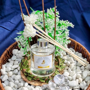 Könnte beinhalten: Ein klarer Glasdiffusor mit silbernem Deckel, beschriftet mit "Blissful Ambience Gift Gallery", steht in einem geflochtenen Korb mit weißen Steinen. Die Anordnung umfasst weiße Blumen, grünes Laub und Schilf-Diffusorstäbchen.