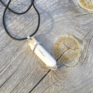 May include: A white crystal pendant with a silver wire wrap, hanging from a black leather cord. The pendant is on a wooden surface.