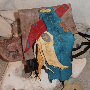 May include: A leather pouch with a flap closure, decorated with colorful leather scraps and stitching. The pouch is made of brown leather and has a blue, red, and yellow leather design. The pouch is closed with a leather strap and buckle.