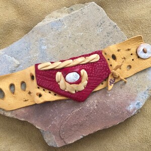 May include: A leather bracelet with a red and tan design. The bracelet has a white stone in the center and a tan leather strap with a button closure.