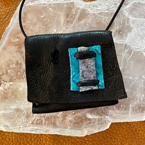 May include: A small black leather pouch with a blue and white patterned flap. The pouch is hanging from a black cord.