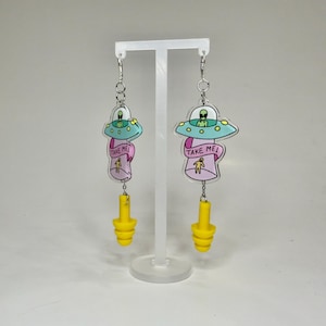 May include: A pair of dangle earrings featuring a clear acrylic charm with a cartoon alien spaceship and the text "Take Me!"  The charm is attached to a yellow silicone earplug.