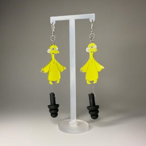 May include: A pair of novelty dangle earrings. Each earring features a yellow cartoon character with a white collar and black earplugs. The earrings hang from silver-coloured hooks and chains. The character has a surprised expression.