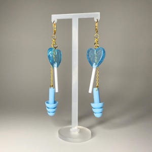 May include: A pair of novelty earrings. Each earring has a blue heart-shaped pendant with glitter, suspended from a gold-coloured chain. Below, a white stick and a light blue earplug-shaped charm. Displayed on a clear stand.
