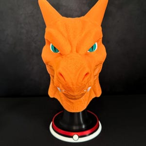 Könnte beinhalten: Eine orangefarbene Charizard-Maske mit türkisfarbenen Augen und weißen Zähnen. Die Maske ist auf einem schwarzen Ständer mit einer rot-weiß-schwarzen Basis montiert, die einem Pokéball ähnelt. Die Maske hat eine strukturierte Oberfläche und ist detailliert.