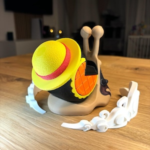 May include: A 3D printed snail with a yellow and red hat, holding a controller. The snail is on a white cloud-shaped stand. The text "Universal Controller Stand - Inspired by OnePiece" is below the stand.
