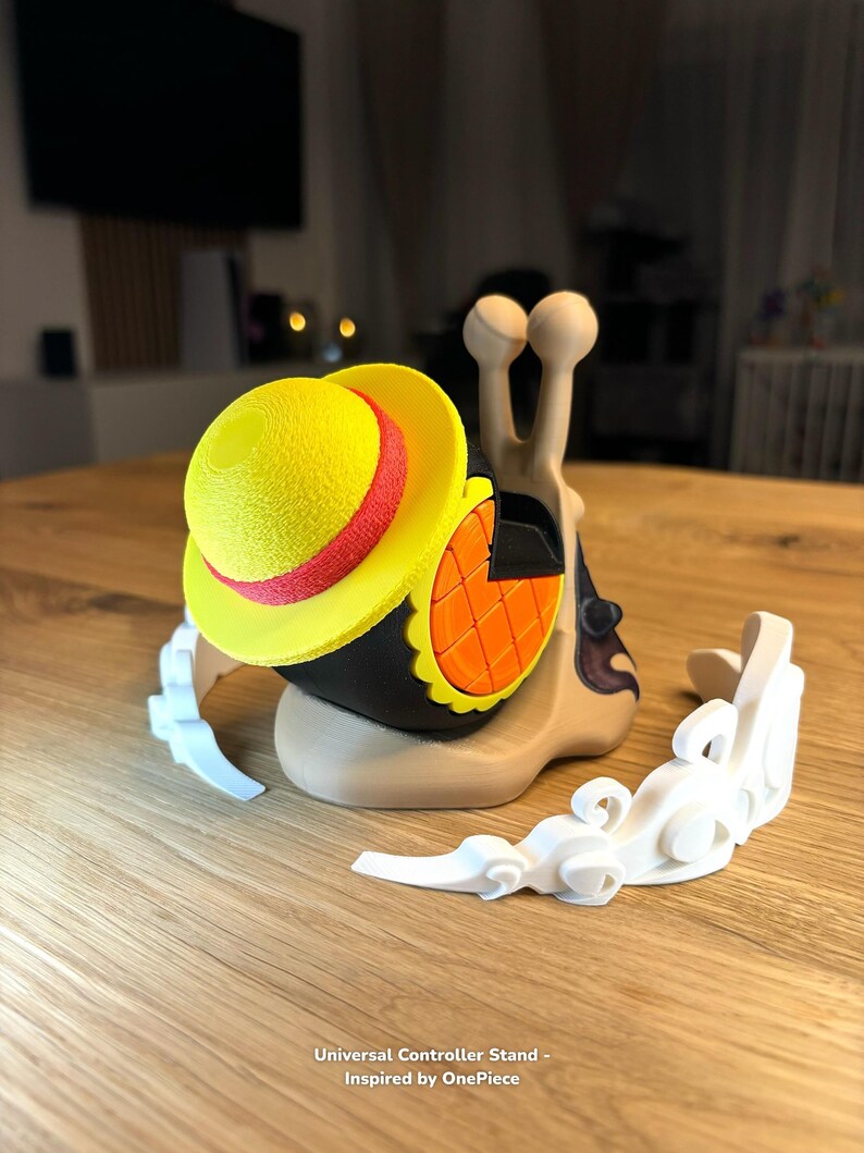 May include: A 3D printed snail with a yellow and red hat, holding a controller. The snail is on a white cloud-shaped stand. The text "Universal Controller Stand - Inspired by OnePiece" is below the stand.