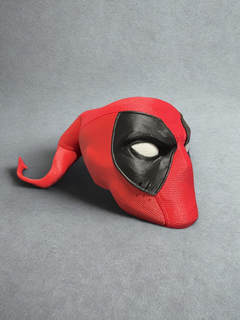 May include: A red and black Deadpool mask. The mask is primarily red with a textured surface and features black eye coverings and a curved, pointed extension on the left side. The eye openings have a white mesh material.