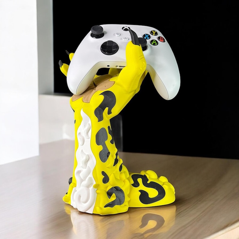 May include: A yellow and black monster hand holding a white video game controller. The monster hand is a stand for the controller.