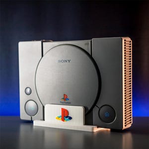 Puede incluir: Una consola Sony PlayStation negra con una unidad de disco gris y una ventilación de color cobre en el lado derecho. La consola está sentada en un soporte blanco con el logotipo de PlayStation.