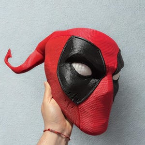 May include: A red Deadpool mask with black eye coverings and white eye lenses. The mask has a textured surface and a curved extension on the left side. The mask is held up against a light blue background.