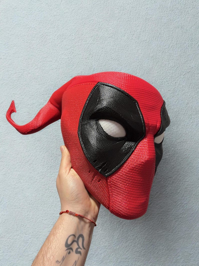 May include: A red Deadpool mask with black eye coverings and white eye lenses. The mask has a textured surface and a curved extension on the left side. The mask is held up against a light blue background.