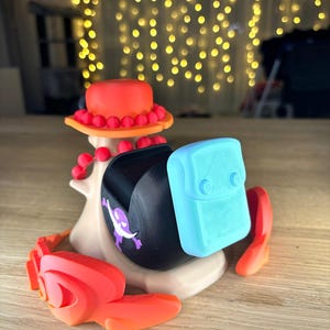 May include: A 3D printed figurine of a character wearing a red and orange hat with a blue and white backpack. The figurine is standing on a wooden surface.