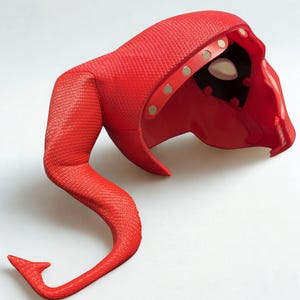 May include: A red, textured mask with a curved tail. The mask has a hood and eye openings. Gold-colored rivets secure the hood. The tail curves upward and ends in a small, pointed tip. The mask is made of a flexible material.