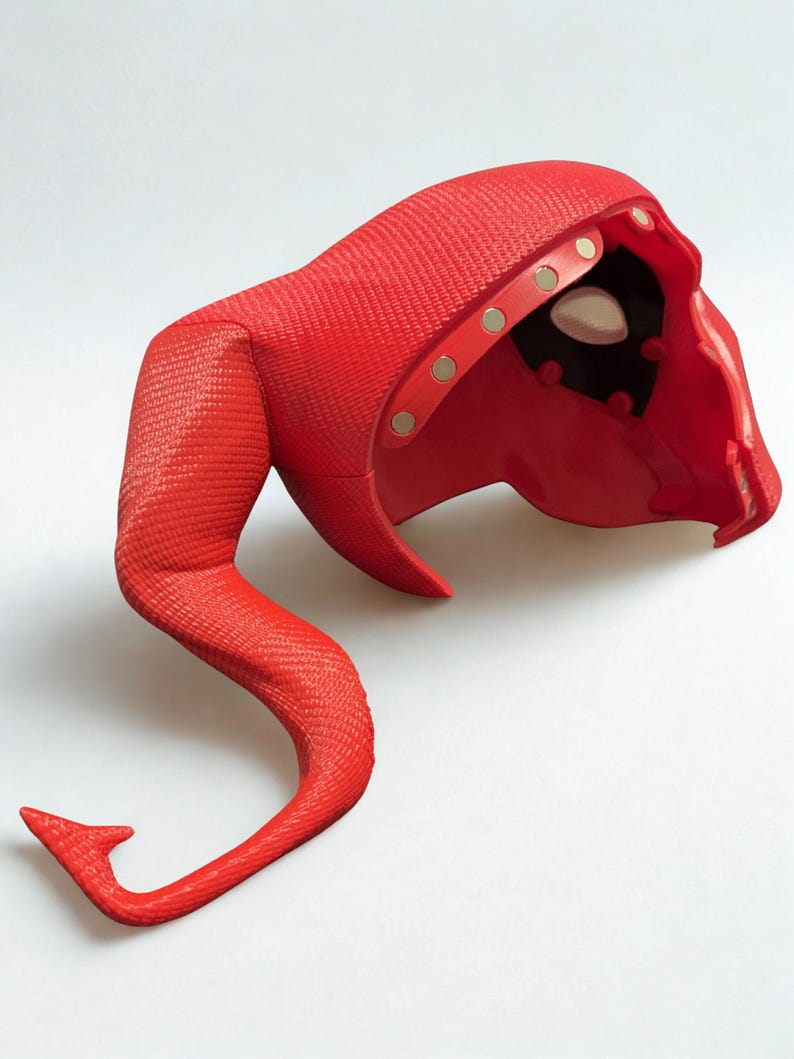 May include: A red, textured mask with a curved tail. The mask has a hood and eye openings. Gold-colored rivets secure the hood. The tail curves upward and ends in a small, pointed tip. The mask is made of a flexible material.
