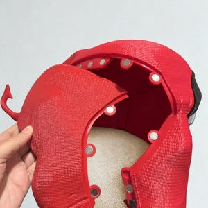 May include: A red, textured mask with a hinged section, revealing the interior. The mask has a detailed design with visible rivets and a small, curved horn-like protrusion. The interior is a lighter color, and the mask appears to be made of a rigid material.