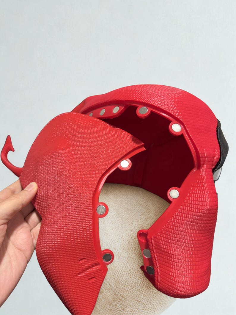 May include: A red, textured mask with a hinged section, revealing the interior. The mask has a detailed design with visible rivets and a small, curved horn-like protrusion. The interior is a lighter color, and the mask appears to be made of a rigid material.