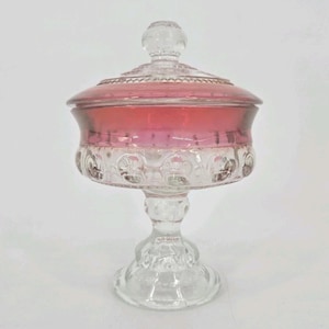 May include: A clear and pink glass candy dish with a lid, featuring a pedestal base and a textured pattern of raised circular motifs around the bowl. The upper portion of the bowl is tinted in a vibrant pink hue, while the lid has a knob handle.