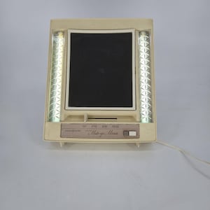 May include: A vintage beige makeup mirror with a rectangular black mirror and two vertical light strips. The mirror has a control panel with settings for day, office, and evening. The text "General Electric" and "Make-up Mirror" are visible.