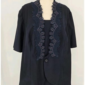 May include: A navy blue two-piece outfit featuring a short-sleeved jacket and a matching top. The jacket has a decorative, dark blue lace trim with beaded accents around the neckline. The outfit is displayed on a mannequin.