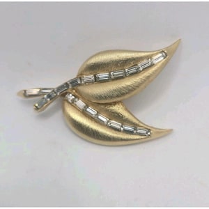 May include: A gold-toned brooch shaped like two leaves with a brushed matte finish. Each leaf features a central vein accented with a row of clear, rectangular baguette-cut stones that extend along the stem of the brooch.