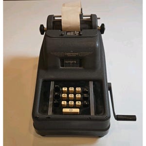 May include: A vintage black Underwood Sundstrand adding machine with a paper roll. The machine has a number pad with white keys and a crank on the side. The text "Underwood Sundstrand" is visible on the front.