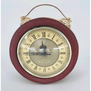 May include: A circular alarm clock featuring a burgundy leather-effect frame and gold-coloured handle and bells. The clock face displays Roman numerals and the word "QUARTZ". The hands indicate approximately 9:15.