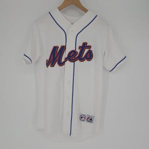 Puede incluir: Camiseta de béisbol blanca con la palabra "Mets" en letras azules y naranjas en el pecho. La camiseta tiene ribetes azules en el cuello, las mangas y la tapeta de botones. Un pequeño parche con el logotipo del equipo es visible en la parte inferior.