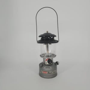 May include: A Coleman Dual Fuel lantern featuring a grey metal fuel tank base, a clear glass globe, and a black metal top vent. The lantern includes a wire carrying handle, a black control knob, and a red and white logo printed on the base.