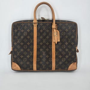 May include: A brown briefcase featuring the iconic dark brown and tan monogram pattern. The bag includes two tan leather top handles, vertical tan leather trim accents, gold-tone hardware, and a matching tan leather luggage tag attached to one handle.