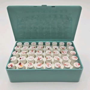 Puede incluir: Una caja de plástico turquesa abierta, llena de numerosos carretes blancos pequeños. Cada carrete tiene un diseño impreso en la parte superior, con varios tonos de rojo y rosa. La caja está sobre una superficie blanca.