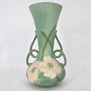 May include: A matte sage green ceramic vase featuring a flared rim and two symmetrical open-work handles. The body is decorated with embossed, light cream-colored dogwood flowers and green stems that extend upward along the neck of the vessel.