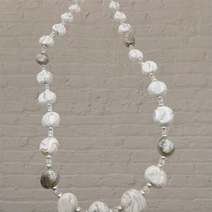 Puede incluir: Un collar de cuentas blancas con un patrón similar al mármol y detalles plateados. El collar presenta una variedad de cuentas redondas, algunas con un acabado brillante. El collar mide aproximadamente 46 cm de largo.