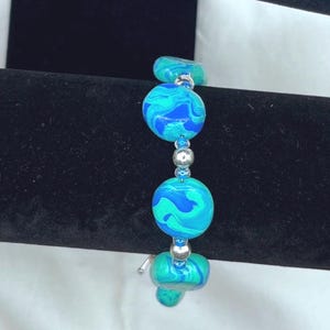May include: A beaded bracelet featuring large, round beads in shades of blue and teal with a marbled effect. The bracelet is strung on a silver-colored chain, creating a striking contrast. The bracelet is a piece of jewelry.