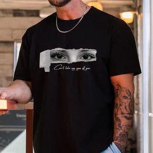 以下が含まれることがあります： 破れた紙の効果で目が描かれた黒いTシャツ。「Can't take my eyes off you」の文字が下にプリントされています。Tシャツはクルーネックと半袖です。男性は水色のズボンを履いています。