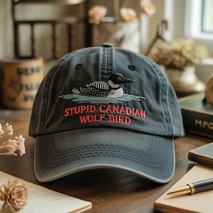 May include: A dark gray baseball cap with a curved brim. The cap features an embroidered design of a loon on water, with the text "STUPID CANADIAN WOLF BIRD" in red. The cap is made of a soft, worn material.