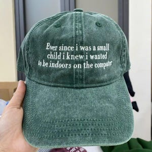Könnte beinhalten: Grüne Baseballkappe mit weiß gesticktem Text: "Ever since I was a small child I knew I wanted to be indoors on the computer." Die Kappe hat einen gebogenen Schirm und ein klassisches Sechs-Panel-Design.