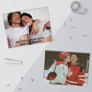 May include: Two spiral-bound calendars featuring photographs of young men. One calendar has the text "HEATED RIVALRY BOYS 2026 CALENDAR". The other calendar shows two men in hockey uniforms. The background is white with office supplies.