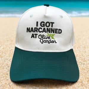 以下が含まれることがあります： 前面に「I GOT NARCANNED AT Olive Garden」と刺繍された白とティールの野球帽。帽子には湾曲したつばと上部にボタンが付いています。背景は砂と海のあるビーチの風景です。
