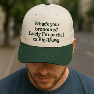 Puede incluir: Gorra de béisbol crema y verde oscuro con el texto "What's your pronouns? Lately I'm partial to Big/Dawg" en negro. La gorra tiene una visera verde oscuro y la lleva una persona. El texto está en una fuente sans-serif.