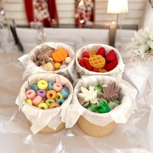 Puede incluir: Cuatro pequeños vasos de papel blanco llenos de fundidos de cera de colores. Los fundidos de cera tienen forma de rosquillas, frutas y hojas. Los vasos están sobre una superficie blanca.