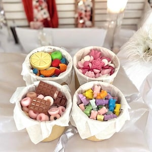 Puede incluir: Cuatro pequeños vasos de papel blanco llenos de fundidos de cera de colores. Los vasos están dispuestos en un cuadrado sobre una superficie blanca. Los fundidos de cera tienen forma de fruta, flores y osos.