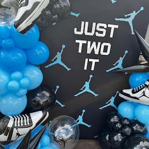 以下が含まれることがあります： 「JUST TWO IT」の文字と、ジャンプする人の水色のシルエットが描かれた黒い背景。 黒と青の風船とスニーカー型の風船で飾られたシーン。
