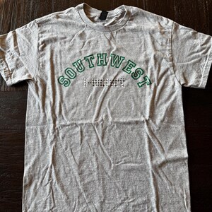 Pode incluir: Camiseta cinza mesclada com a palavra "SOUTHWEST" em verde arqueada sobre o peito. Abaixo, a palavra "T-BIRDS" é exibida em um padrão preto e branco. A camiseta é feita de material macio.