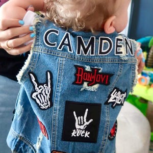 Puede incluir: Un chaleco vaquero azul con parches, incluyendo "CAMDEN", "Bon Jovi", "Guns N' Roses" y "Rock" en la espalda.