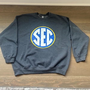 May include: Dark gray sweatshirt with the letters "SEC" in white, inside a blue circle with a yellow border. The sweatshirt is laid flat on a wooden surface.