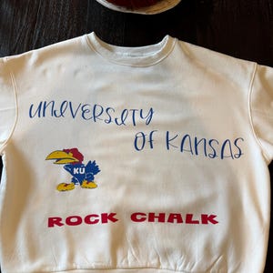May include: Cream-colored sweatshirt with the text "UNIVERSITY OF KANSAS" in blue script. Below the text is a cartoon Jayhawk mascot and the words "ROCK CHALK" in red block letters. The sweatshirt has a crew neck.