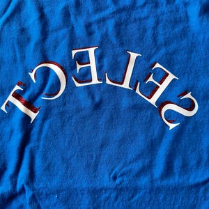 May include: A royal blue t-shirt with the word "SELECT" arched across the front in white letters with red glitter accents. The shirt is made of a soft, slightly wrinkled fabric.