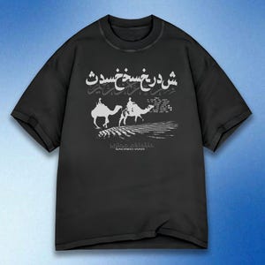 Può includere: T-shirt nero con un design grafico bianco raffigurante due cammelli e testo arabo. Il design include le parole "Sacred War" in basso. La maglietta ha maniche corte e girocollo.
